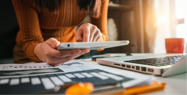 Person looks at iPad while leaning over desk full of laptop and papers with graphs shown